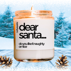 A white candle with a label that says "Dear Santa... do you like it naughty or nice" is placed on a snowy surface with pine cones in the background.